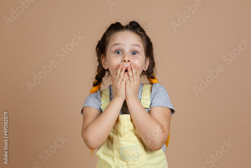 Portrait of shocked excited little girl with kanekalon braids, closing mouth with hands making grimace looking at camera wearing yellow jumpsuit and gray t-shirt on beige background.