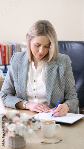 left handed woman boss writing in a notebook planning working day. employer woman wearing formal chothes in office working in the morning. vertical shot