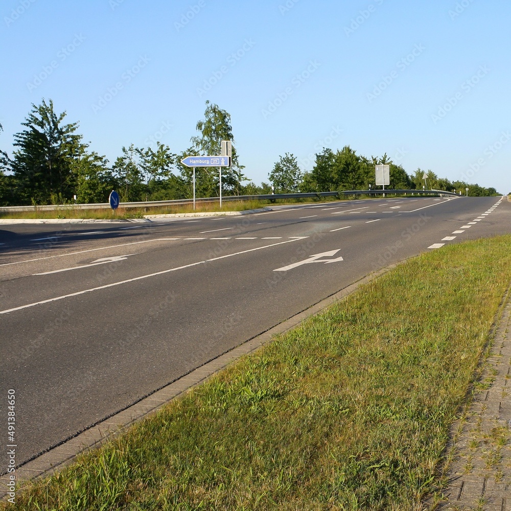 Fototapeta premium View of empty road in Itzehoe, Germany with traffic direction sign to Hamburg and trees in clear blue sky background. No people.
