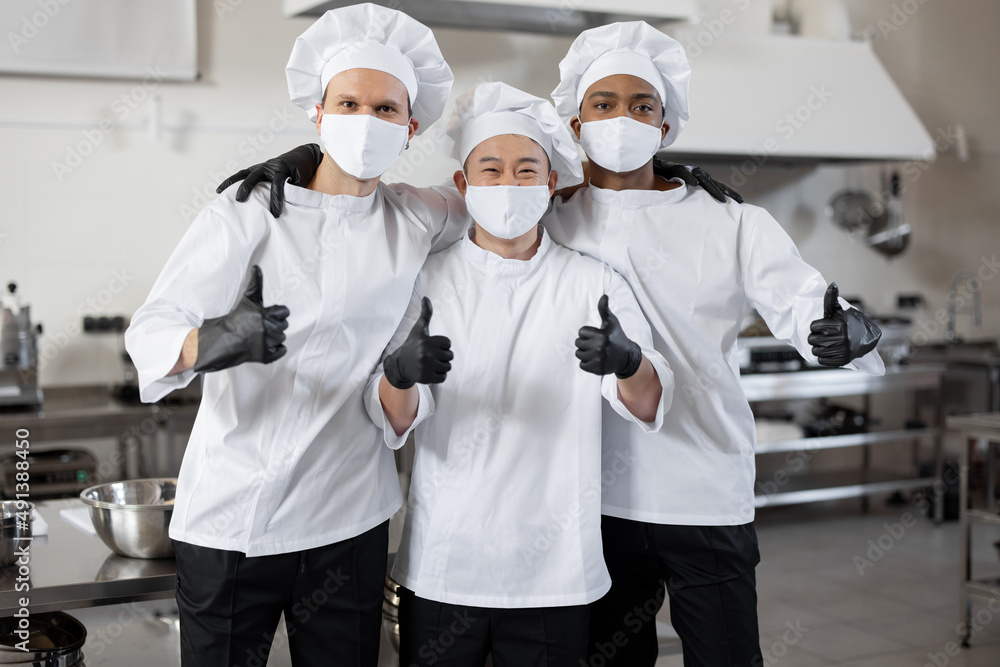 Portrait of multiracial team of three chefs standing together and showing OK sign. Well-dressed chefs wearing face masks and protective gloves to prevent infection spread during pandemic