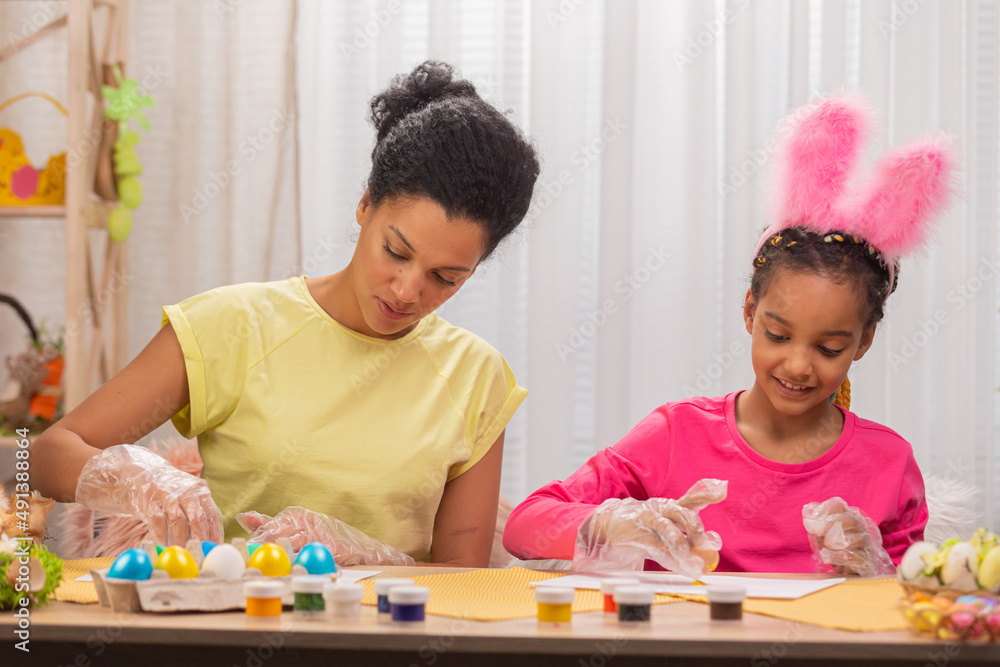 Mom and daughter paint eggs using yellow paint. African American woman and little girl are sitting in decorated room at home. Family is preparing for spring holiday. Happy easter. Close up. Mom and daughter paint eggs using yellow paint. African American woman and little girl are sitting in decorated room at home. Family is preparing for spring holiday. Happy easter. Close up.