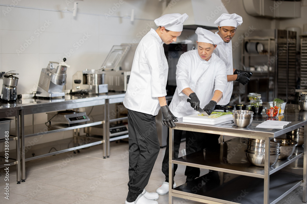 Three multiracial chefs having some conversation while cooking in professional kitchen. Interior with professional equipment and stainless tables. Teamwork at restaurant