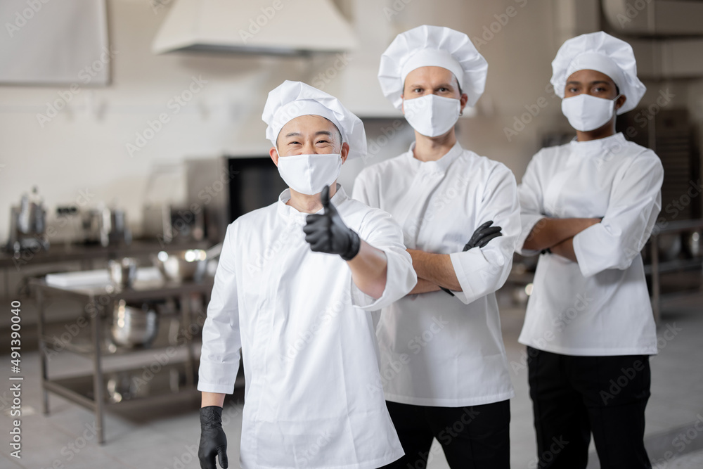Portrait of multiracial team of three chefs standing together in the professional kitchen. Well-dressed chefs in face masks and protective gloves ready for a job. New normal for business during