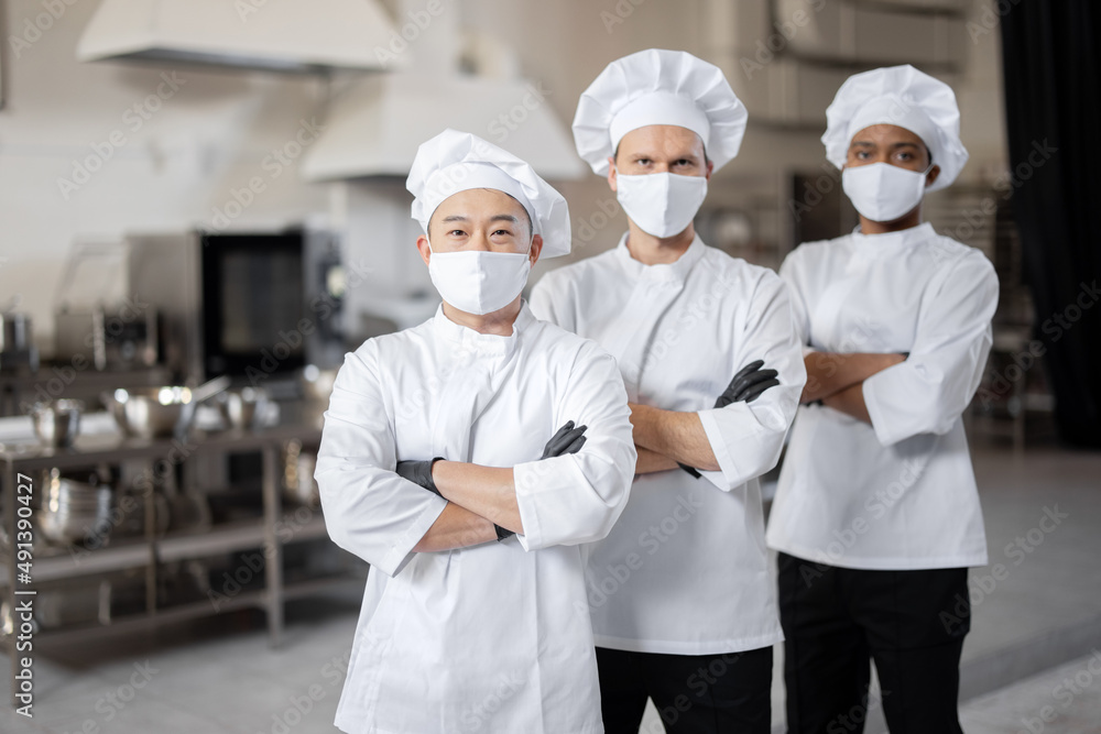 Portrait of multiracial team of three chefs standing together in the professional kitchen. Well-dressed chefs in face masks and protective gloves ready for a job. New normal for business during