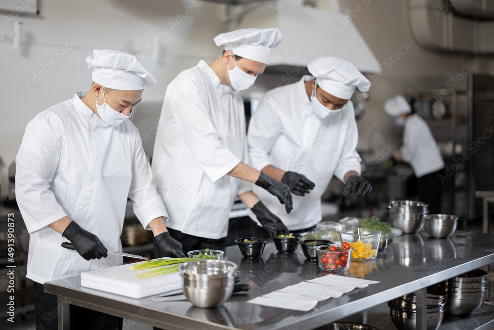 Three well dressed chefs in face masks prepare takeaway food in professional kitchen. Concept of a dark kitchen for cooking for delivery during pandemic Three well dressed chefs in face masks prepare takeaway food in professional kitchen. Concept of a dark kitchen for cooking for delivery during pandemic
