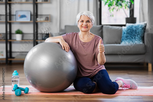 sport, fitness and healthy lifestyle concept - smiling senior woman with exercise ball showing thumbs up sitting on mat at home