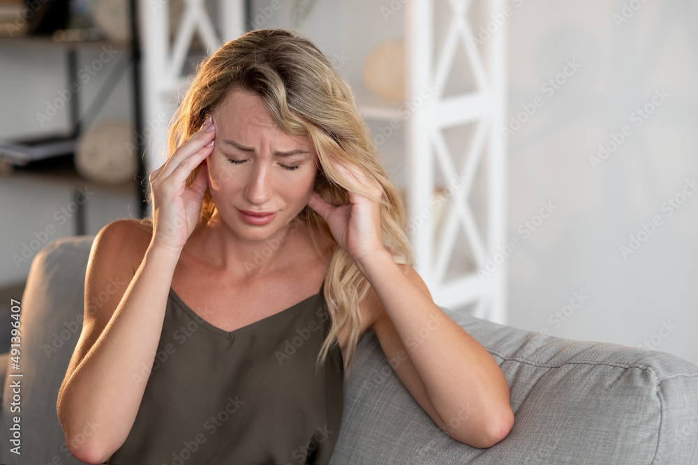 Exhausted day. Suffering woman. Headache frustration. Feeling bad sick lady holding massaging head temples sitting sofa light room interior.