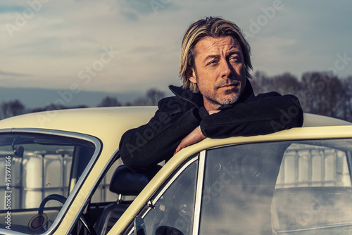 Man in a black coat with blond hair and stubble stands by a classic car in sunny countryside. #491397866