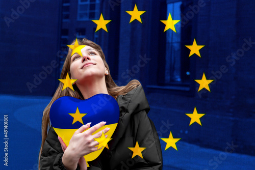 War in Ukraine. Young cute girl holding a yellow-blue heart-shaped balloon against the background of the flag of the European Union (EU). Concept of Ukraine's accession to the European Union (EU).
