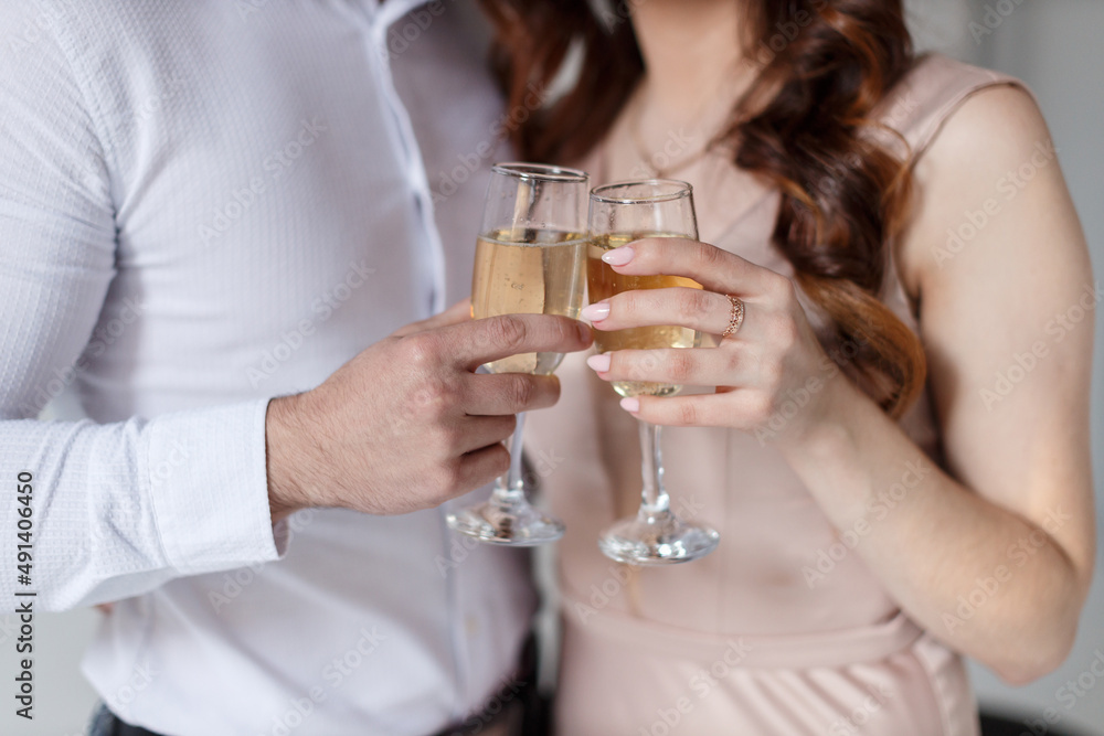 A young man in a white shirt and a girl with a ring on his hand in a beige dress drink champagne in crystal glasses, close-up without faces A young man in a white shirt and a girl with a ring on his hand in a beige dress drink champagne in crystal glasses, close-up without faces