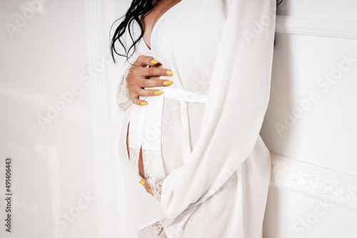 A pregnant thin woman with a yellow manicure holds a big belly with her hands in a transparent white peignoir on a white background, close-up without faces
