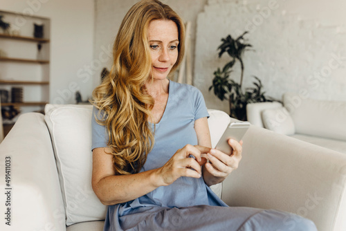 Portrait of blond long-haired woman in her living room using cell phone, surfing web and reading messages, news, updating applications and mobile software. People and technology concept