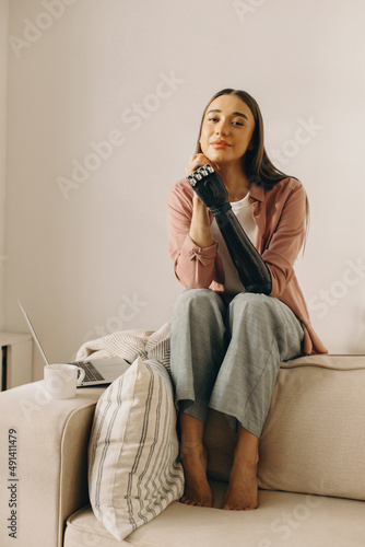 Pretty charming woman with robotic black mechanical prosthesis instead of amputated arm sitting on back of white couch next to opened laptop and cup of tea, looking at camera with dreamy romantic look