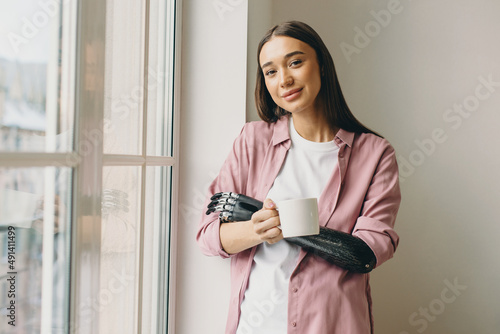 Attractive beautiful youthful brunette girl with bionic black prosthetic hand holding mug of coffee standing next to huge white window, admiring beauty of morning, looking at camera with cute smile