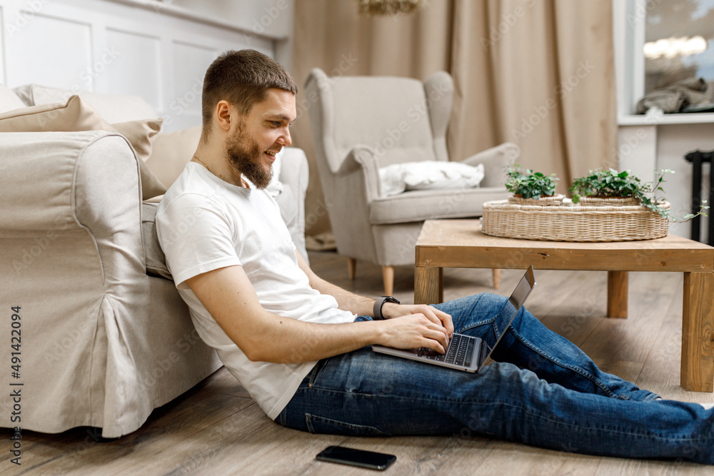 young man at home on the floor near the sofa working on a laptop with a phone.