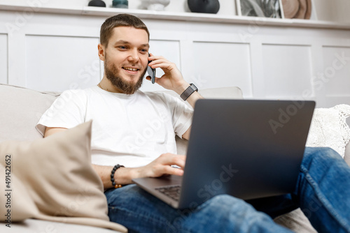 young man at home on the couch talking on the phone and looking at the laptop.