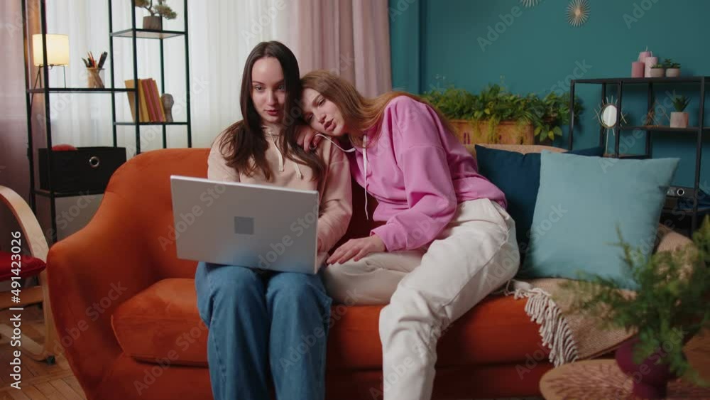 Happy girls friends siblings using laptop computer watching online tv show video sitting on sofa together. Young adult women couple looking at notebook screen making online shopping purchase at home Happy girls friends siblings using laptop computer watching online tv show video sitting on sofa together. Young adult women couple looking at notebook screen making online shopping purchase at home