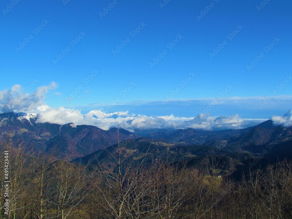 Scenic view of forest covered landscape of pre-alpine Slovenia with the peaks of the mountains in the Kamnik-Savinja alps out of the clouds on a clear blue day from Porezen in Gorenjska, Slovenia