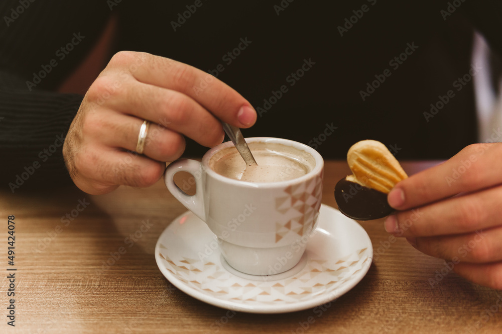 Hands of an unrecognizable young man with a cup of coffee and a cookie in the foreground. Hands of an unrecognizable young man with a cup of coffee and a cookie in the foreground.
