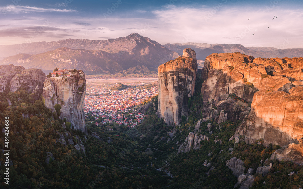 Scenic panoramic view of the Monastery of the Holy Trinity in Meteora at sunrise and Kalambaka town at the distance. Tourist and pilgrimage experience. Natural and religious wonders of Greece.