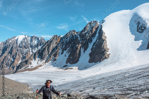 Hiker with trekking poles walks near large glacier tongue in high mountains. Scenic landscape with tourist near glacier in sunlight. Man with sunglasses and camera among snow mountains in sunny day.