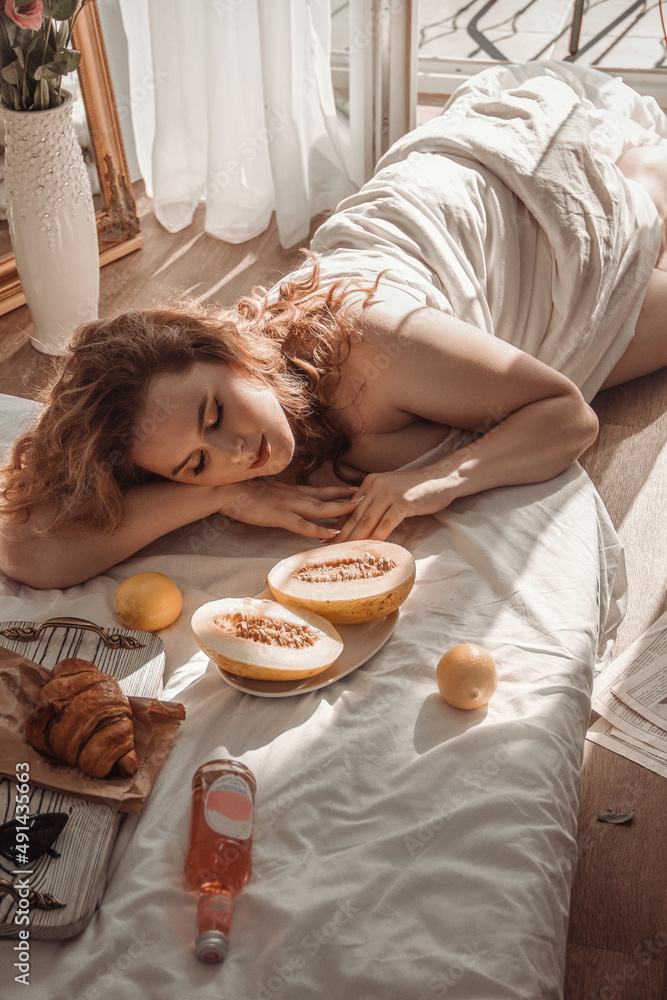 young girl plus size with red curly hair is lying near half melons and  croissant with lemons and smiling cute on the white bed background, camera from top. lifestyle morning concept, free space