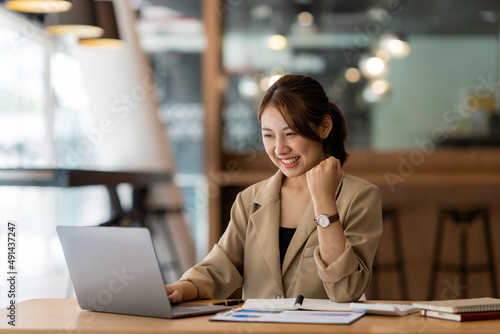 Happy of Asian young businesswoman see a successful business plan on the laptop computer, pen on wooden table background in office, business expressed confidence embolden
