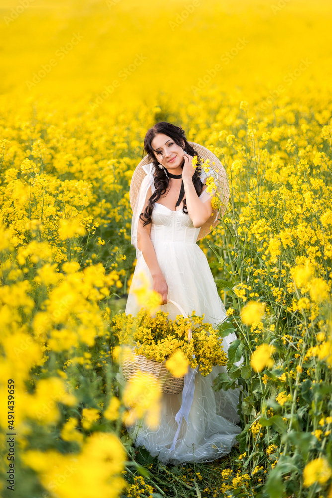 young beautiful woman in a field with a large basket of yellow flowers. A girl in a long white dress collects rapeseed young beautiful woman in a field with a large basket of yellow flowers. A girl in a long white dress collects rapeseed