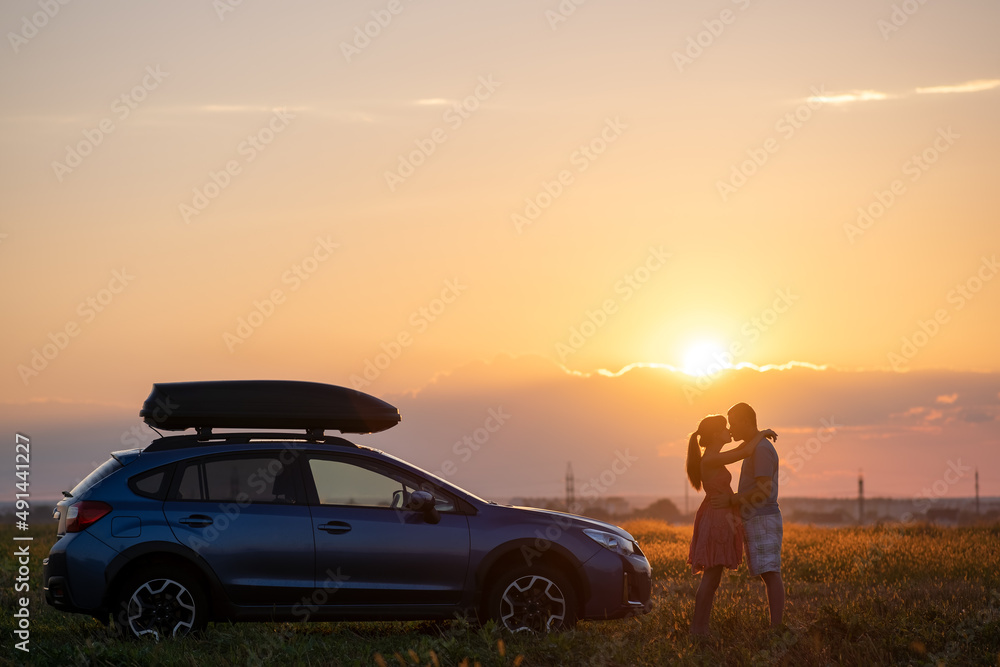 Happy couple spending time together near their SUV car during honeymoon road trip at warm summer evening. Young man and woman enjoying road trip travelling by vehicle in nature