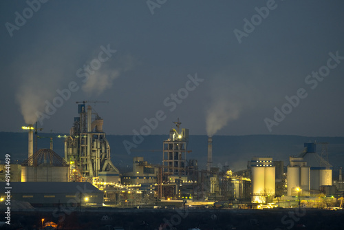 Illuminated cement plant with high factory structure and tower cranes at industrial production area at night. Manufacture and global industry concept © bilanol