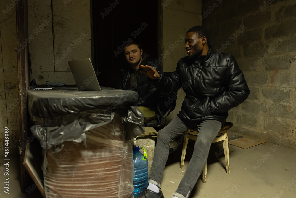 Caucasian guy and black guy are hiding in a basement during a bombardment and having a talk near laptop Caucasian guy and black guy are hiding in a basement during a bombardment and having a talk near laptop