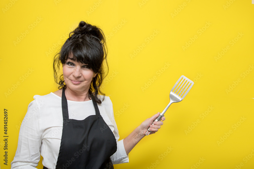 beautiful aged lady cook in black apron with cooking spatula pointing gesture on yellow background, concept offer, advertising, copy space beautiful aged lady cook in black apron with cooking spatula pointing gesture on yellow background, concept offer, advertising, copy space
