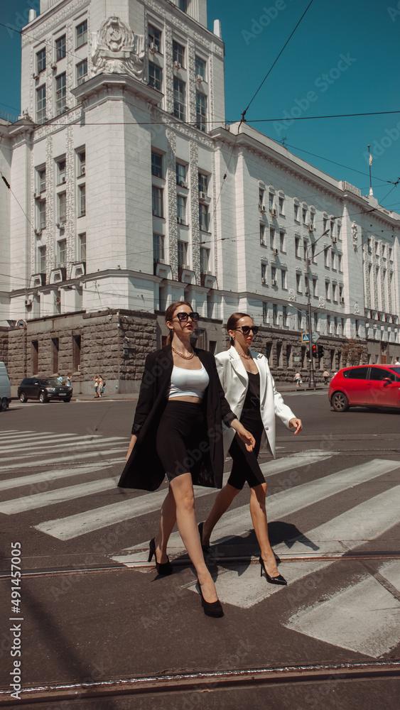 two sports young girls in suits and sunglasses are walking fashion along the crosswalk on the street modern building background. fashion concept, free space