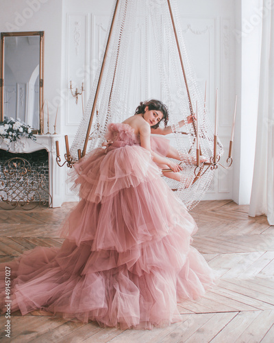 young brunette girl in pink princess dress with train is sitting on the crystal chandelier near panoramic window and mirror on a white wall background like dior style. fashion concept, free space photo