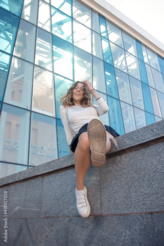 young curly brunette teen girl in white sweater and black skirt is sitting outdoor with leg in camera near blue glass building background and looking happy with smile. lifestyle concept, free space