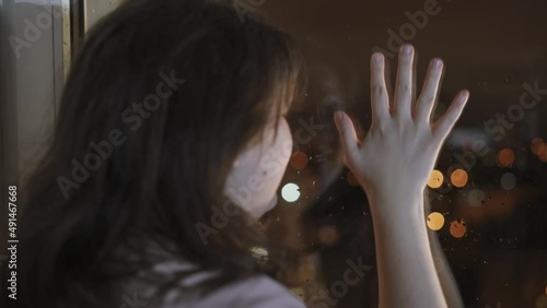 Sad young woman knocks on window with fist, runs fingers down glass with drops, leaving marks. Girl in depression and despair screams, reflection of her face on glass. Night in city, light shine.