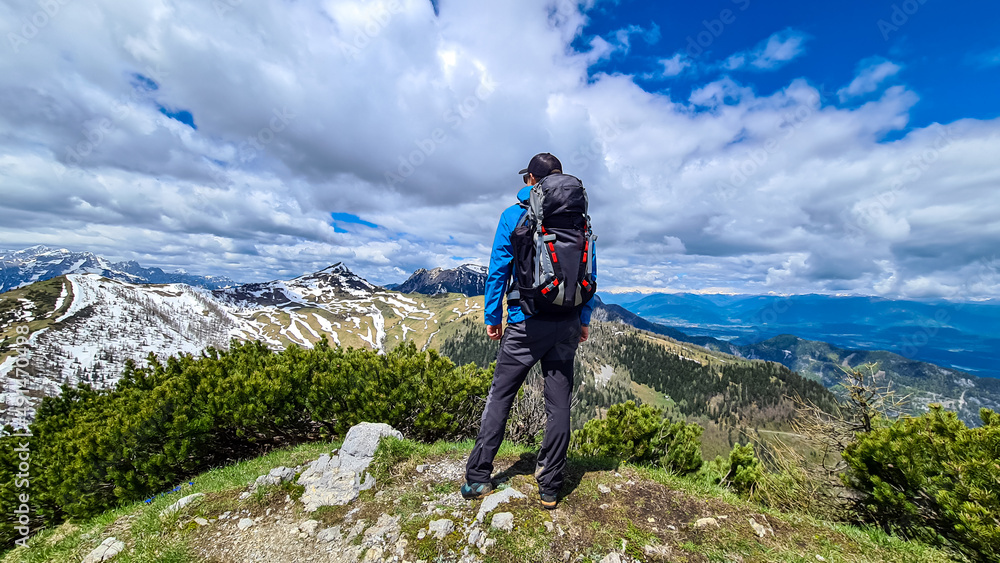 Hiker man with backpack at the summit of Hahnkogel (Klek) with scenic view on mountain peaks in the Karawanks and Julian Alps, Carinthia, Austria. Border with Slovenia. Triglav National Park