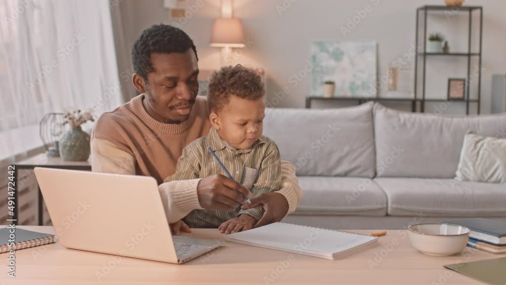 Medium slowmo of Black man paying attention to his toddler son while working on laptop from home during lockdown Medium slowmo of Black man paying attention to his toddler son while working on laptop from home during lockdown
