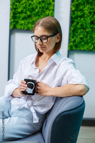 A young woman in glasses with a smile on her face, she is in a white shirt and jeans sitting on a sofa with a cup of tea
