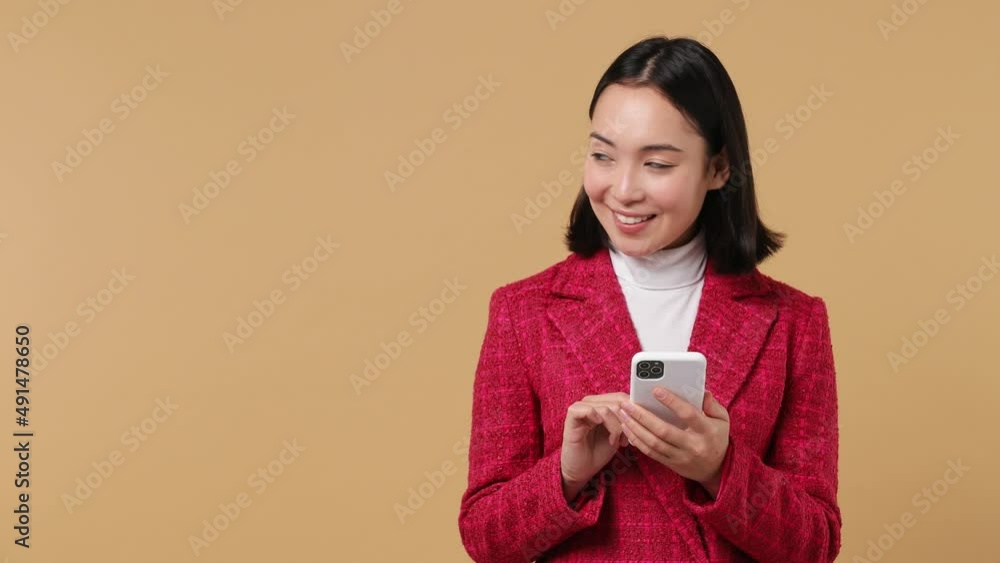 Young woman of Asian ethnicity 20s wears red jacket hold use mobile cell phone look aside on workspace copy space mockup promo commercial area isolated on plain pastel beige background studio portrait