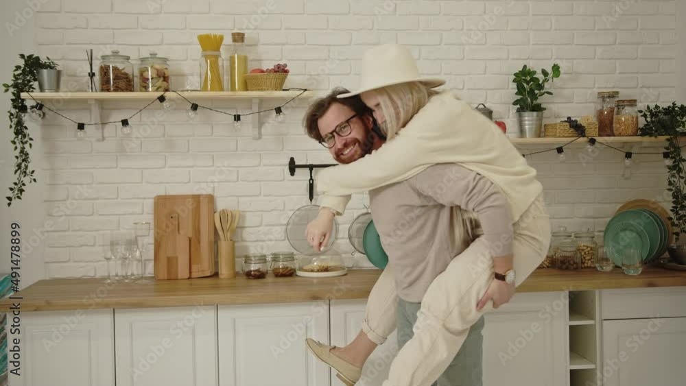 European middle-aged bearded man having fun and lifting his fiancee piggyback in their modern bright kitchen. High quality 4k footage European middle-aged bearded man having fun and lifting his fiancee piggyback in their modern bright kitchen. High quality 4k footage