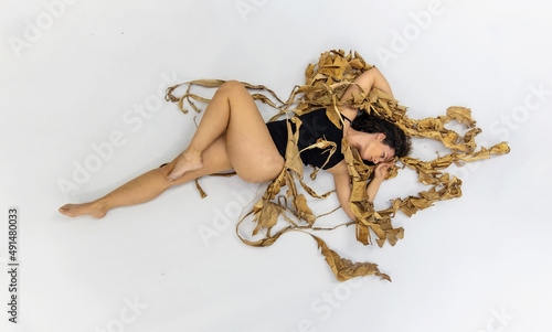 sexy woman with dark brown hair in black dress, covered with dry, withered decorative banana tree leaves lying on the studio floor photo