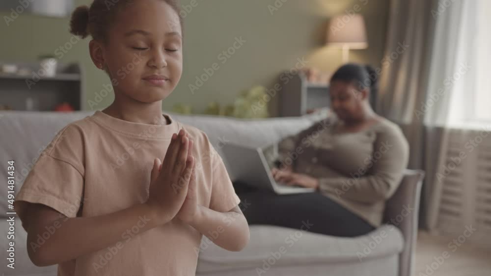 Medium slowmo of 5 year old African-American girl meditating in living room and her mother using laptop while working from home in background Medium slowmo of 5 year old African-American girl meditating in living room and her mother using laptop while working from home in background