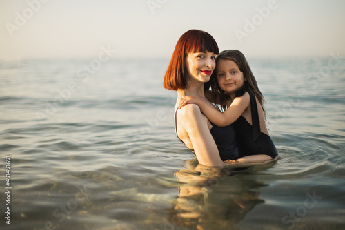 Happy mother with red hair holding daughter in a black swimming suit in the sea. Happy beautiful mother and daughter in swimsuits swimming in sea, family look concept.