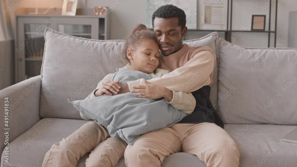 Medium slowmo of African American man and his pretty 8 year old daughter sitting together on couch embracing each other, staying at home during lockdown Medium slowmo of African American man and his pretty 8 year old daughter sitting together on couch embracing each other, staying at home during lockdown