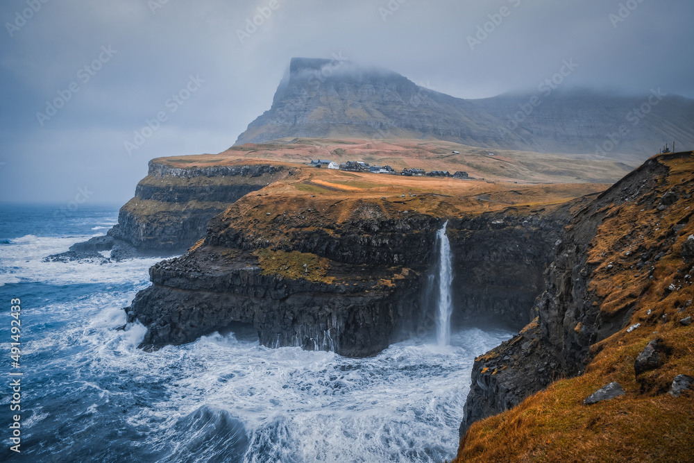 Dramatic view of Mulafossur waterfall with Gasadalur village on background. Vagar island, Faroe Islands, Denmark. November 2021.