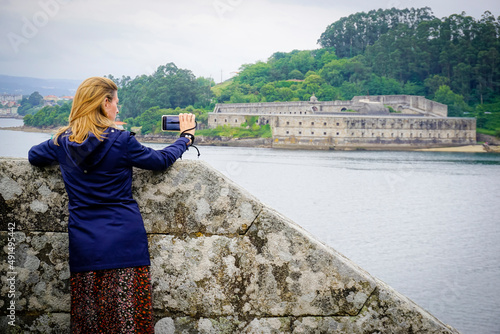 blonde young woman with blue coat taking photos at Castillo de San Felipe. Stone and earth coastal castle from the 16th century with views of the Ferrol estuary photo