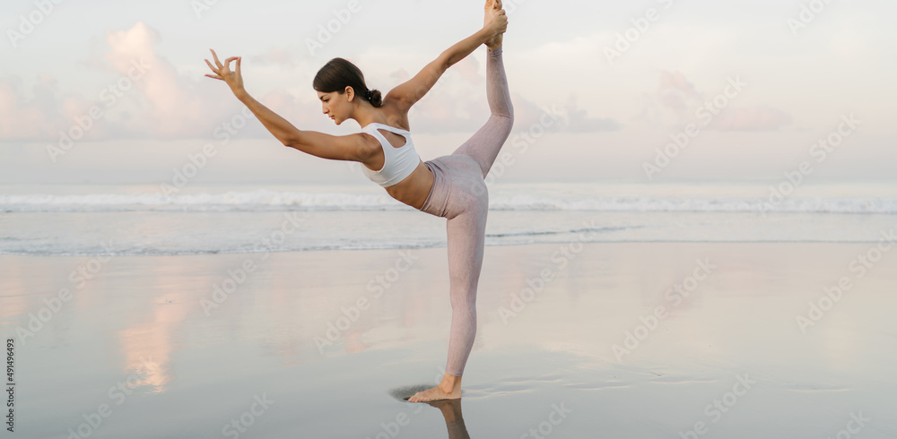Obraz premium Caucaisan woman with good body shape concentrated on hatha asana pose with raised leg, young female in sportive tracksuit stretching for feeling vitality during holistic healing on seashore retreat