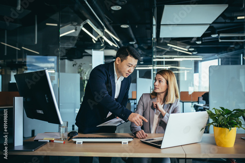 two business people consulting on a project looking at computer monitor screen sitting in the office. Asian male employee Coworker explains colleagues ideas and strategies accountant, financial report