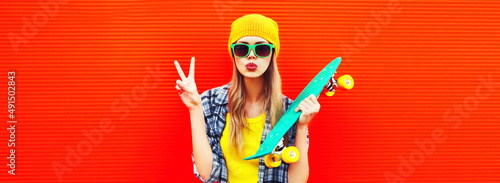 Portrait of stylish blonde young woman model with skateboard wearing colorful yellow hat on red background
